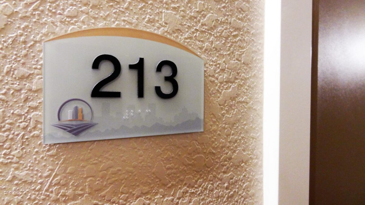 A room sign on a textured beige wall displays the number "20713" in large, black print. Below the number is Braille text, and in the bottom left corner, there is a small illustration of a cityscape. The sign is rectangular with a curved top.