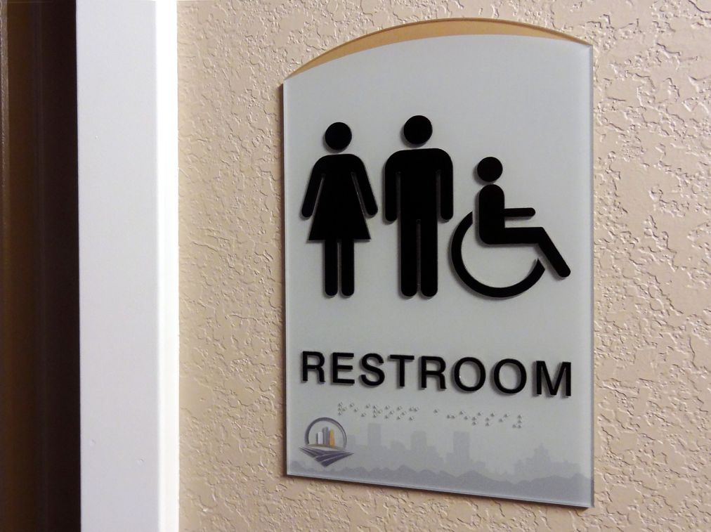 A restroom sign mounted on a wall indicates an accessible bathroom for all genders. The sign shows icons for a woman, a man, and a person in a wheelchair above the word "Restroom." A small logo is visible at the bottom of the sign.