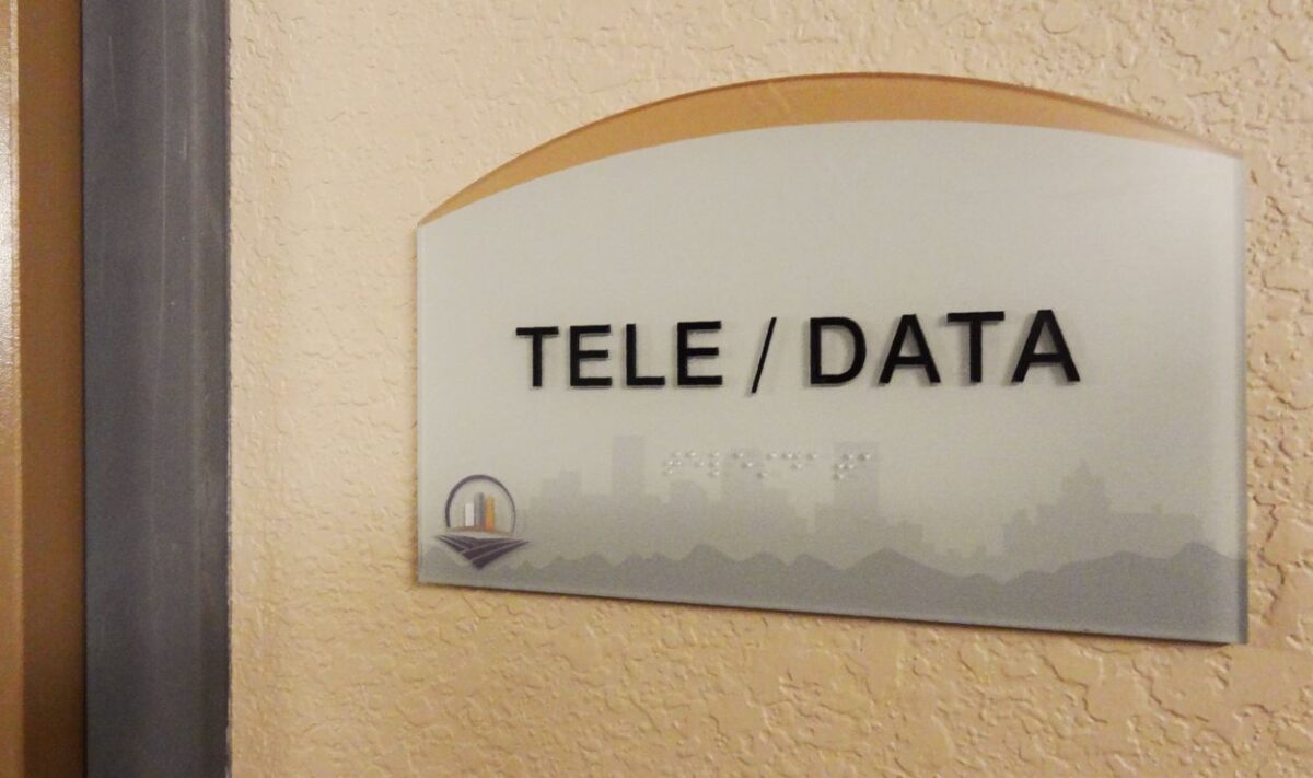 A frosted glass sign on a beige wall reads "TELE / DATA" in bold black letters. Beneath the text, embossed but barely visible, it says “Utilities.” The bottom of the sign features a small, colorful logo.