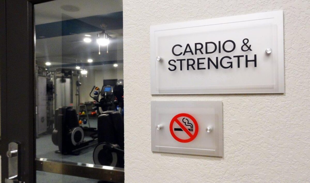 A door with a sign that reads "Cardio & Strength" leads to a gym. Below it, there's a "no smoking" sign. Inside the gym, exercise equipment is visible including a treadmill and elliptical machines. The gym area is lit with ceiling lights.