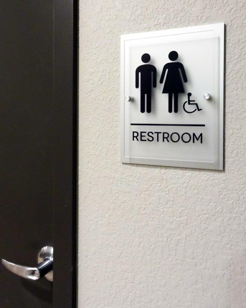 A sign on a wall indicates a restroom. The sign has symbols for a man, a woman, and a person in a wheelchair. The symbols are in black on a white background above the word "RESTROOM." A door with a metallic handle is adjacent to the sign.
