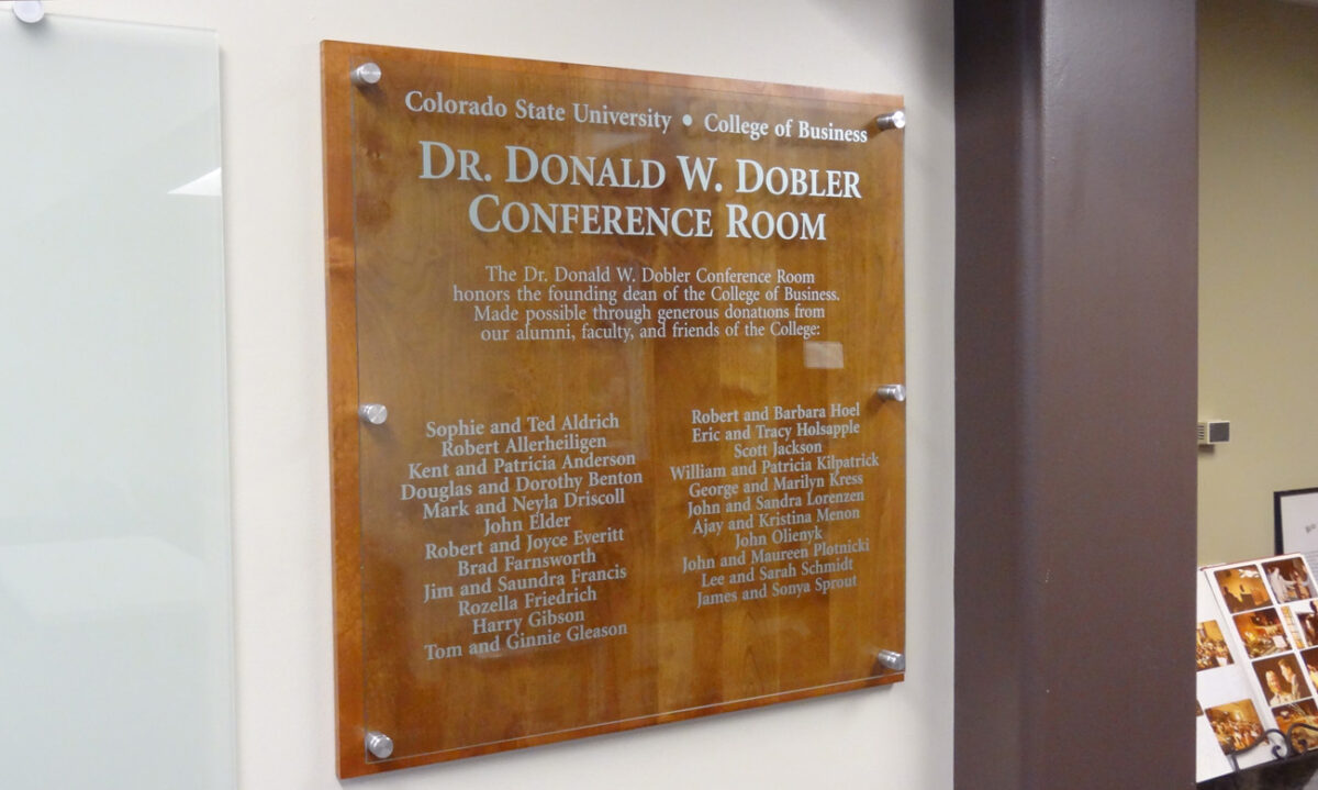 A wooden plaque on the wall reads, "Colorado State University College of Business Dr. Donald W. Dobler Conference Room." The plaque lists names of individuals and organizations beneath the title, honoring their contributions. The plaque is affixed with four metal caps.