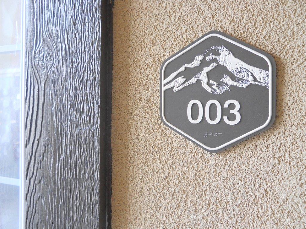 A hexagonal sign on a textured beige wall shows the number "003" with a white mountain graphic above it. To the left, a doorframe with a wood grain pattern is partially visible.
