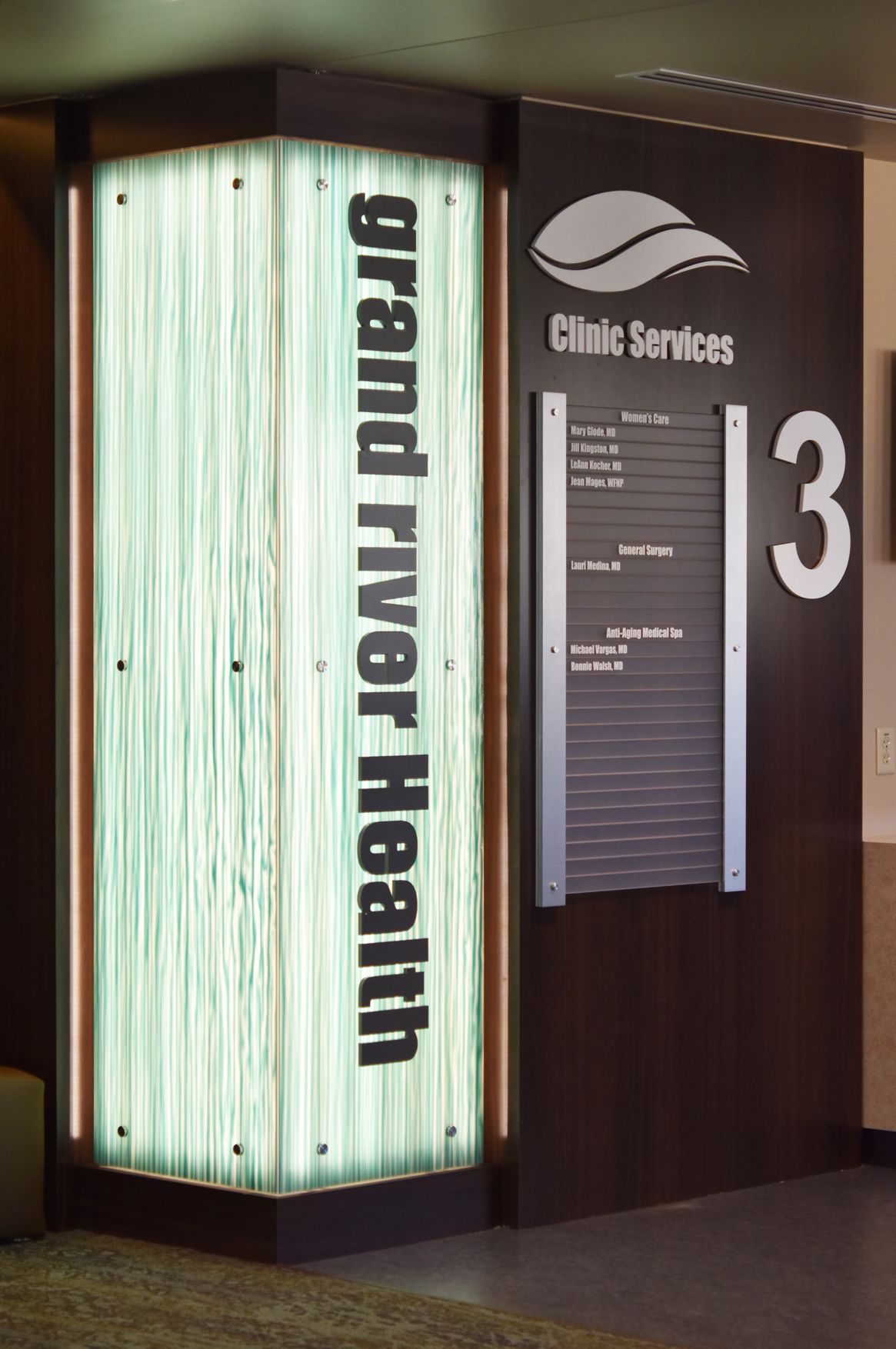 The image shows an illuminated sign for "Grand River Health" next to a directory board for "Clinic Services." The directory lists various departments and room numbers, with a large number "3" to the right of the sign. The decor appears modern and sleek.