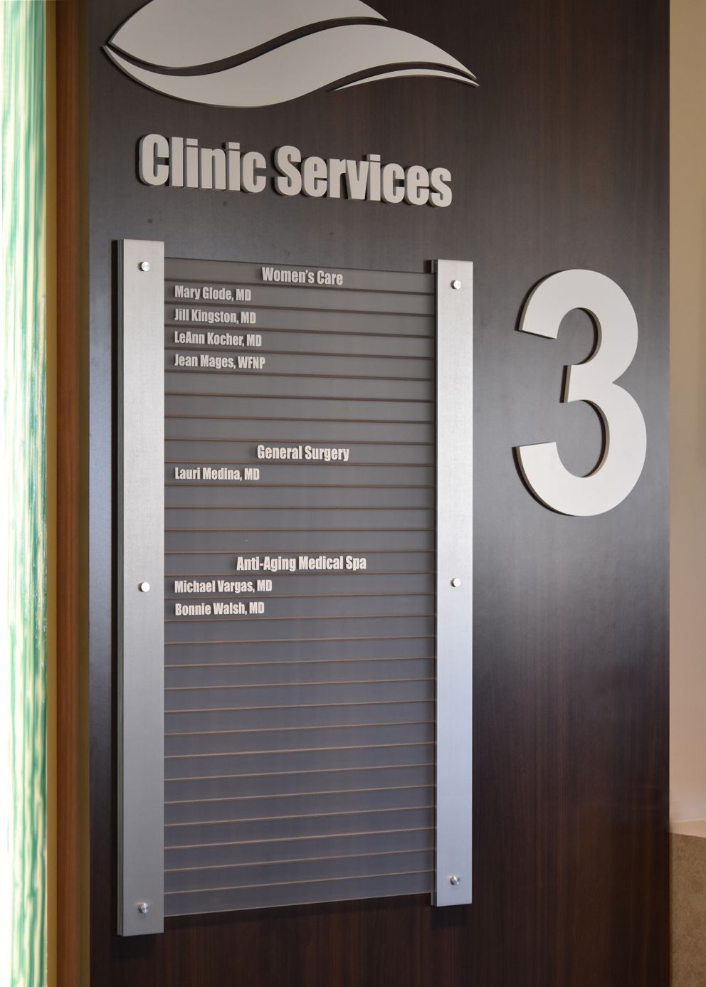 A wall directory sign for Clinic Services on the third floor. It lists Women's Care, General Surgery, and Anti-Aging Medical Spa with various names of doctors alongside each category. The sign is mounted on a dark wooden background.