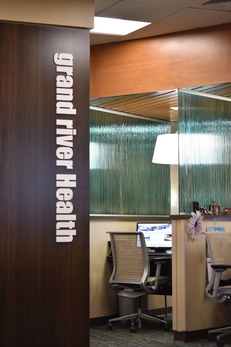 A modern office workspace inside Grand River Health. The office features a wooden partition with "grand river Health" written vertically in white letters. Behind the partition, there are desks with computers and chairs, and a glass wall with vertical lines.