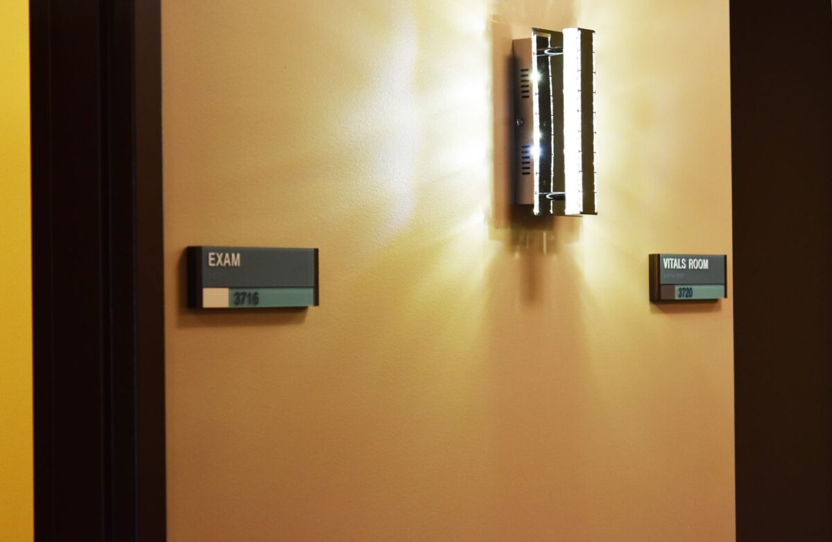 A beige wall with a modern wall sconce emitting light. Two signs are visible: one on the left reads "EXIT" in uppercase letters, and the one on the right reads "ELEVATOR" in uppercase letters. Both signs are green with white text.