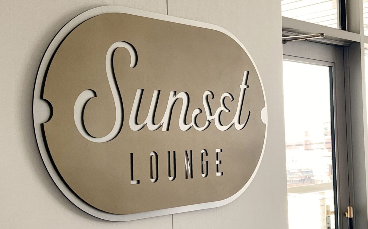 A metallic sign on a wall reads "Sunset Lounge" in elegant script. The background of the sign is a reflective silver color. To the right, there is a window or door with glass panes, allowing light to enter the space.