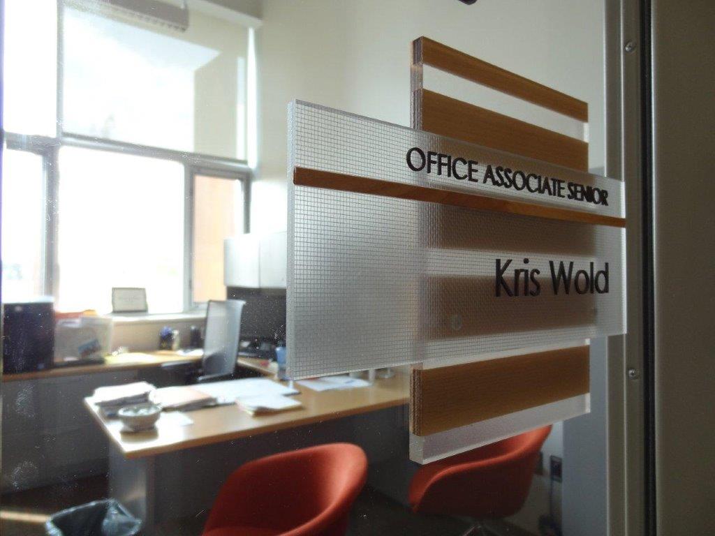 Office door with a nameplate reading "Office Associate Senior, Kris Wold." The door is partially open, revealing a well-lit office with a desk cluttered with papers and a laptop, and two red chairs in front of it. The room appears bright with sunlight coming through windows.
