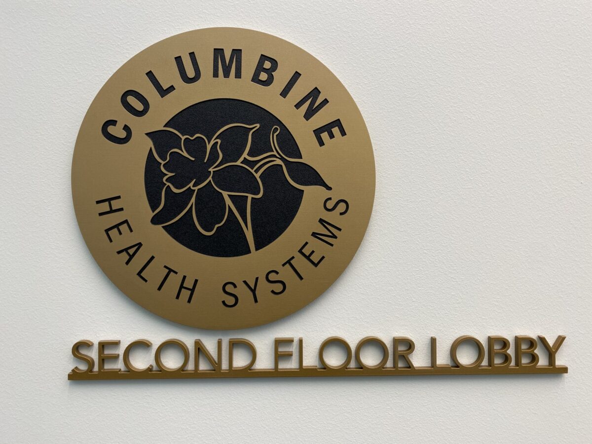 A circular plaque displays "Columbine Health Systems" with a flower logo in the center, mounted on a wall. Below the plaque, "Second Floor Lobby" is written in raised letters.