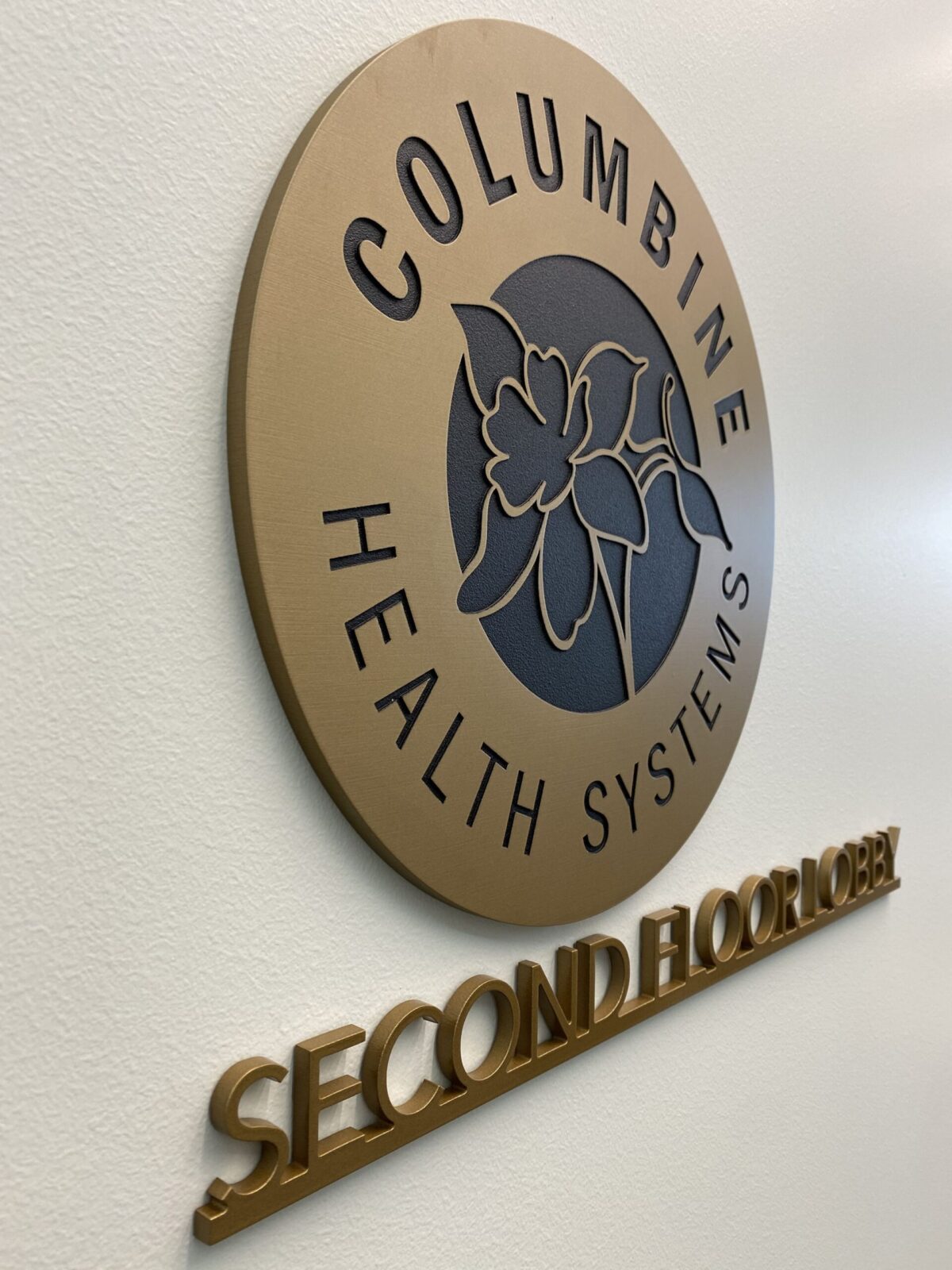 A circular plaque displays "Columbine Health Systems" with a flower logo in the center, mounted on a wall. Below the plaque, "Second Floor Lobby" is written in raised letters.