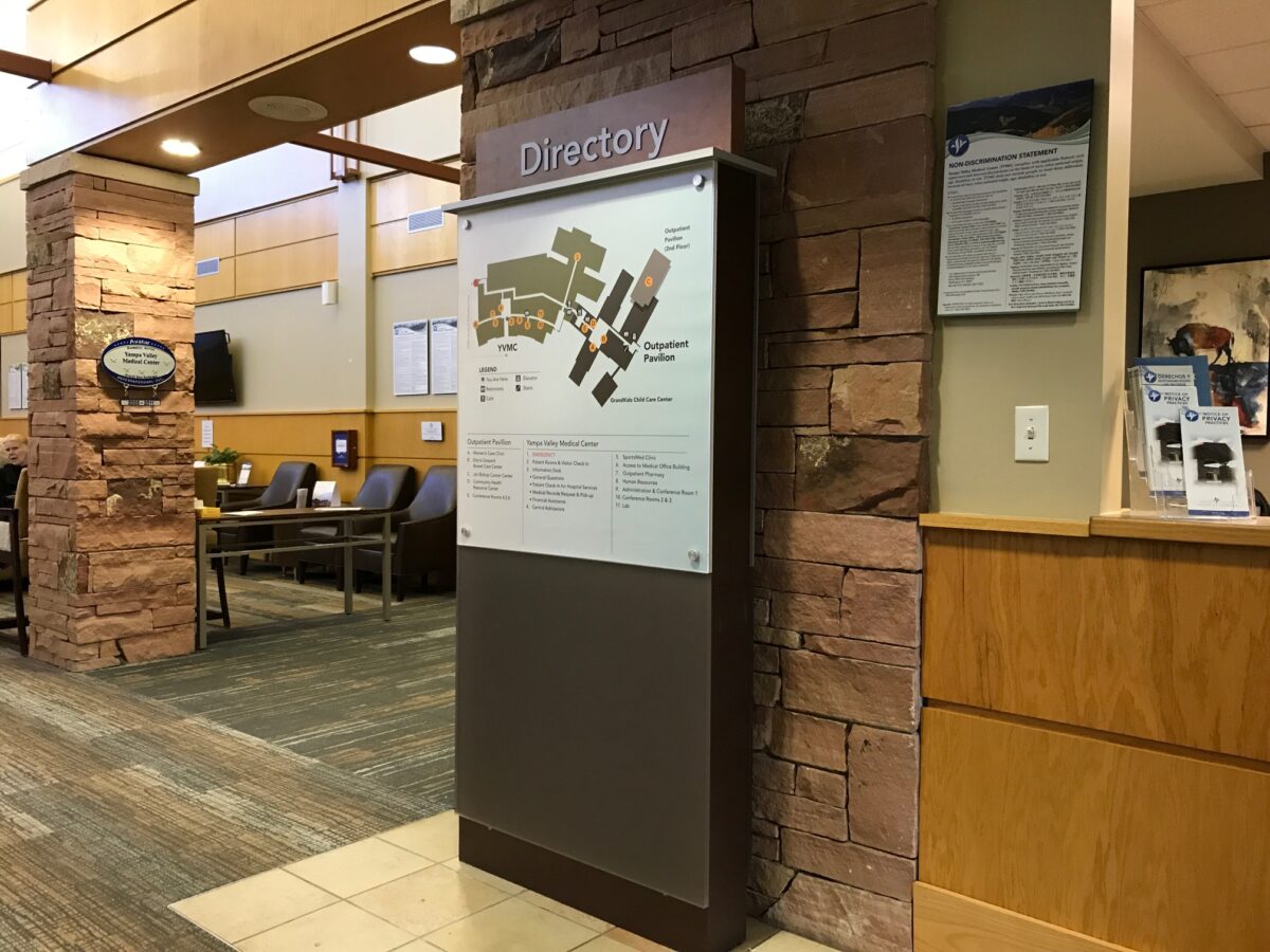 A hospital lobby with stone accents and wooden trim. A large directory sign with a detailed map is prominently displayed. In the background are seating areas with chairs and tables, and a wall-mounted information board is visible on the right.