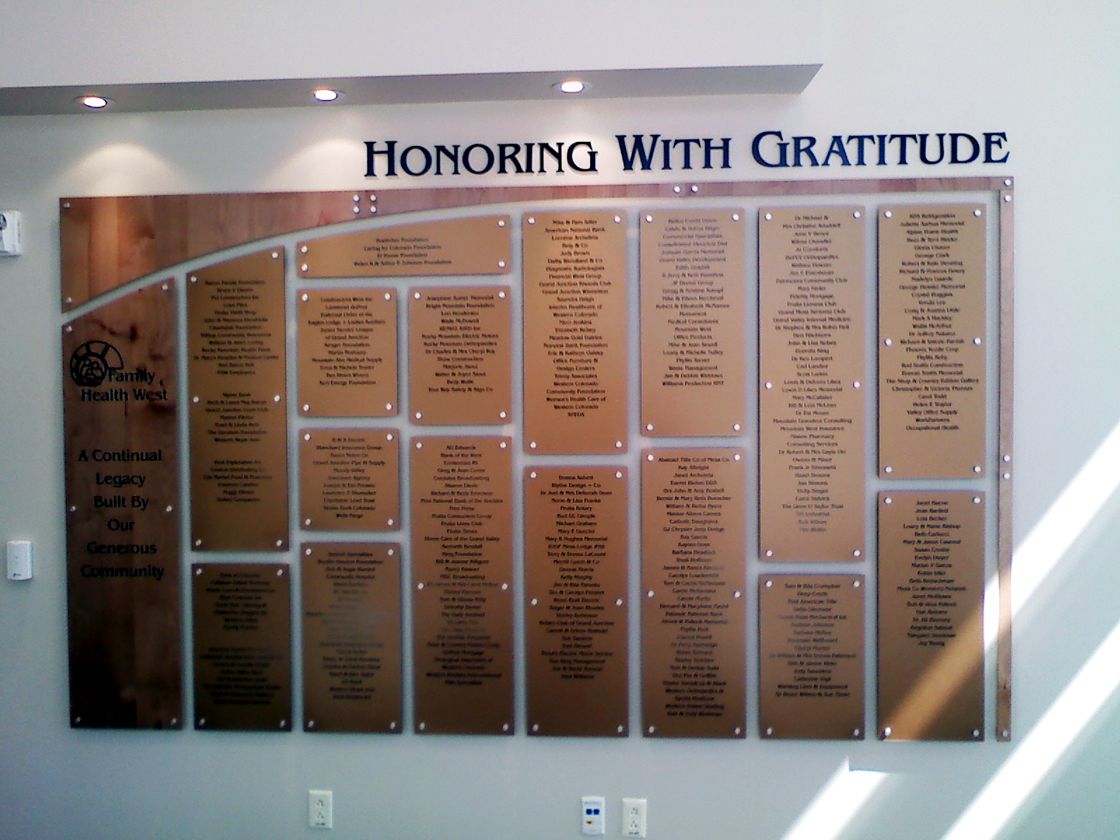 A wall plaque titled "Honoring With Gratitude" displays multiple nameplates with engraved text. The nameplates are arranged in rows and columns on a large board, recognizing individuals for their contributions or achievements.