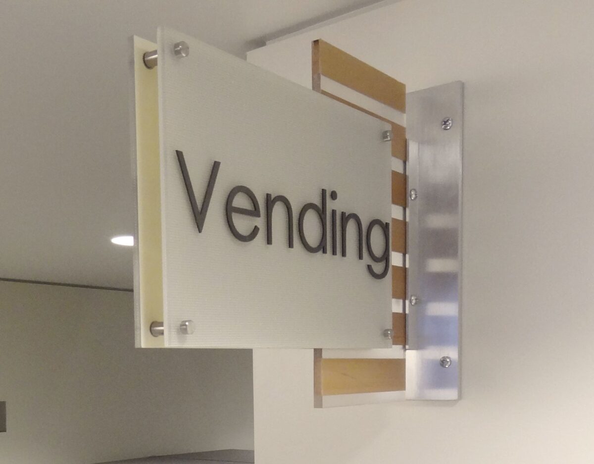A sign mounted on a wall reads "Vending" in black letters on a frosted glass panel, indicating the location of vending machines. The sign is attached to a metal bracket and is lit from above.