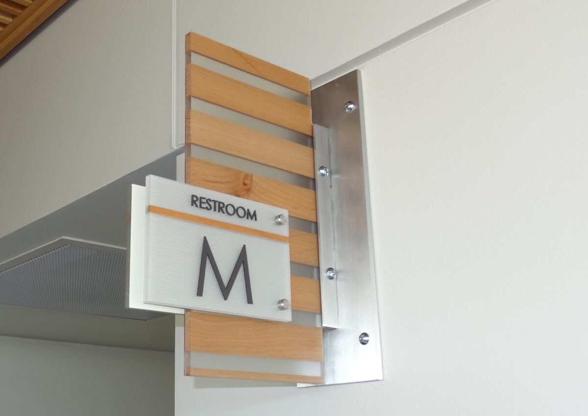 A restroom sign mounted on a wall indicates the men's restroom with a letter "M". The sign is attached to a wooden panel with horizontal slats. The word "RESTROOM" appears above the "M" on the sign.