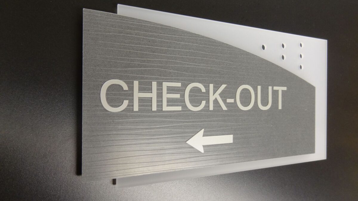 A rectangular sign with a brushed metal finish reads "CHECK-OUT" in large, white letters, with an arrow pointing left beneath the text. The sign is mounted on a dark background.