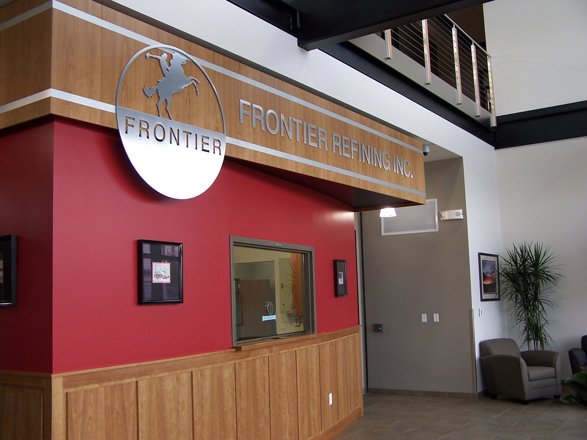 A reception area with a wooden-fronted counter below a sign that reads "Frontier Refining Inc." The interior features a red accent wall, wall-mounted pictures, a potted plant, and a seating area with chairs and tables. The design has modern and rustic elements.
