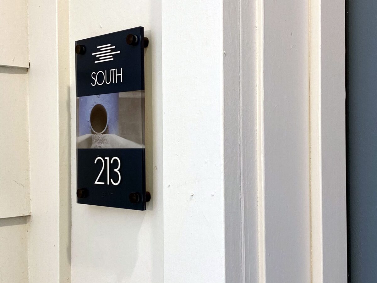 A modern door sign is mounted on a light-colored wall. The sign is black with white text and reads "SOUTH" above a clear section, which has a small circular hole in the center. Below the clear section, the number "213" is displayed in large white letters.