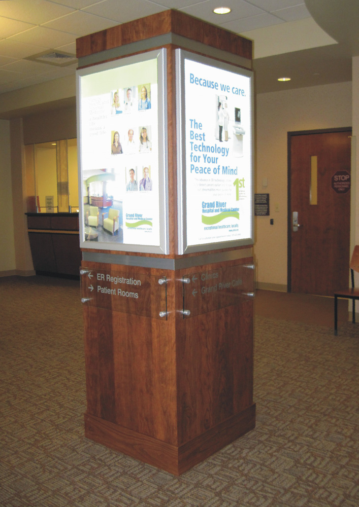 A wooden kiosk stands in a carpeted area with four brightly lit panels displaying various health-related information, images of medical equipment, and texts promoting healthcare services. A reception desk and a door are visible in the background.