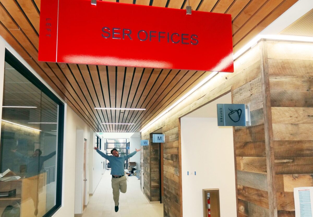 A person mid-jump in a hallway under a red sign that reads "SER Offices." The hallway features wooden paneling on the right side and glass walls on the left. The person is dressed casually and appears to be celebrating or excited.