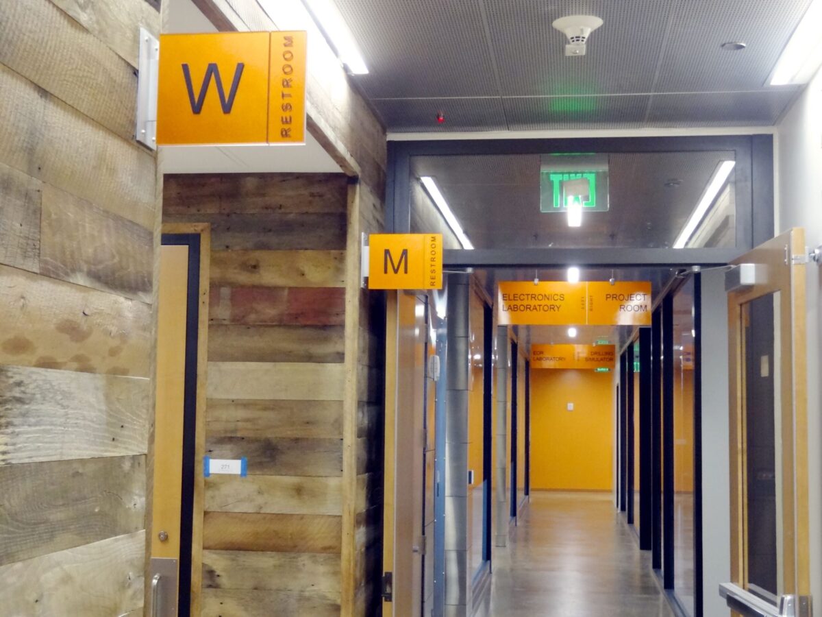 A hallway with wood-paneled walls and orange signs indicating restrooms. One sign with a 'W' for women's restroom is near a door on the left, and another sign with an 'M' for men's restroom is further down the hall. The ceiling has fluorescent lights and an exit sign.