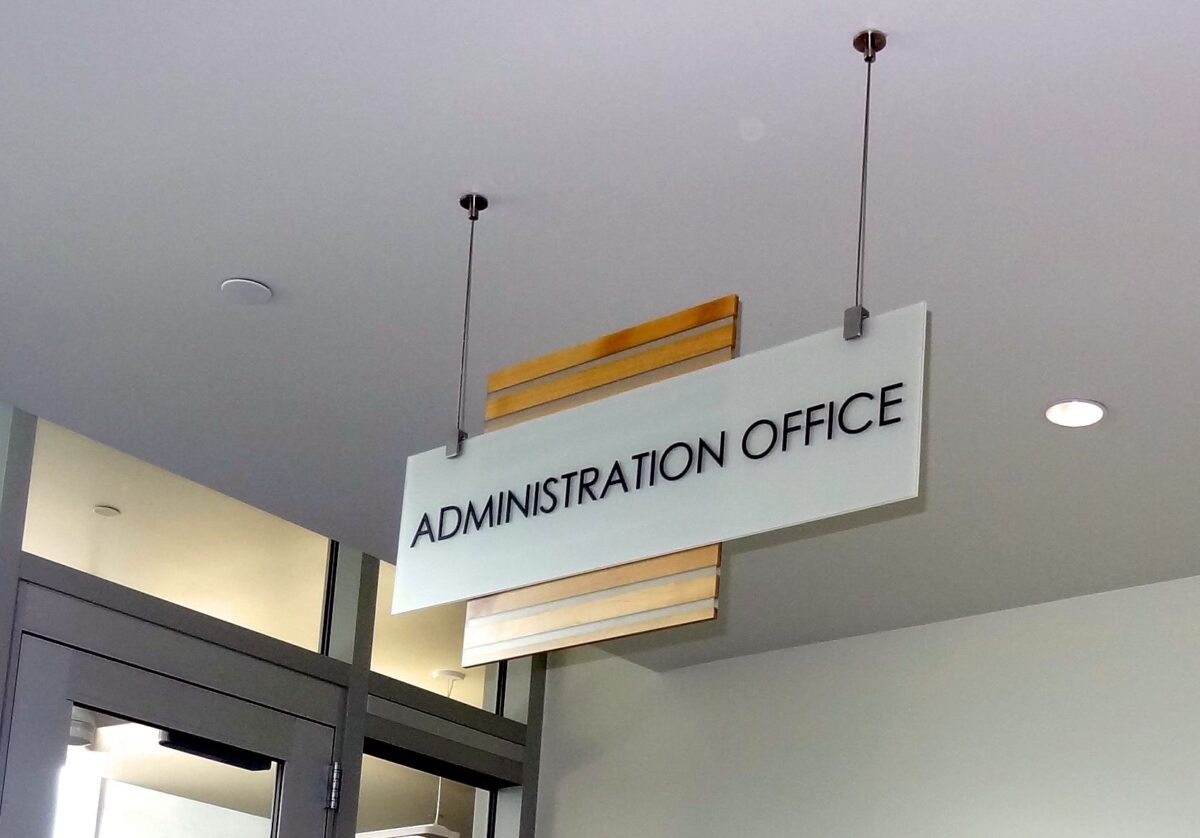 A white rectangular sign with black text reads "ADMINISTRATION OFFICE." The sign is suspended from the ceiling by two wires. The background shows part of a ceiling, walls, and a doorway with glass panels.