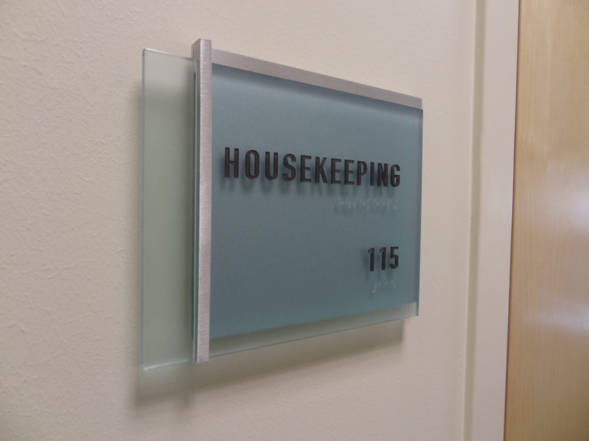 A wall-mounted sign with a frosted glass design and silver trim, displaying the text "HOUSEKEEPING" in bold black letters, and the room number "115" beneath it. The sign indicates the housekeeping office location.