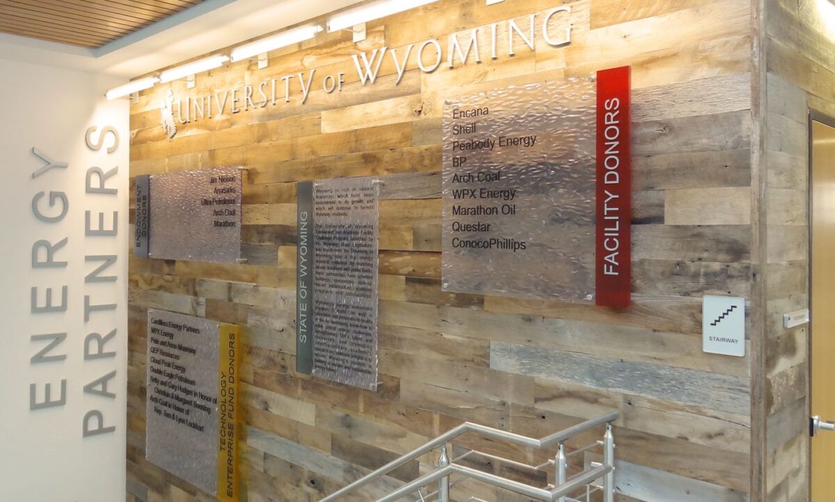 A wooden wall with plaques at the University of Wyoming, showcasing Energy Partners and Facility Donors. The wall is well-lit with spotlights illuminating the text and plaques. There is also a metal railing along the bottom right corner of the image.