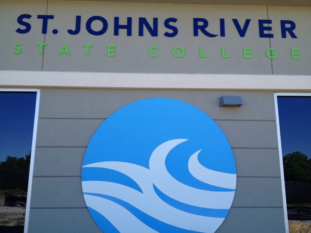 Sign for St. Johns River State College with a blue wave logo against a gray wall. The text "ST. JOHNS RIVER" is in blue, "STATE COLLEGE" is in light green. The background features trees and blue sky partially visible through glass windows on either side.