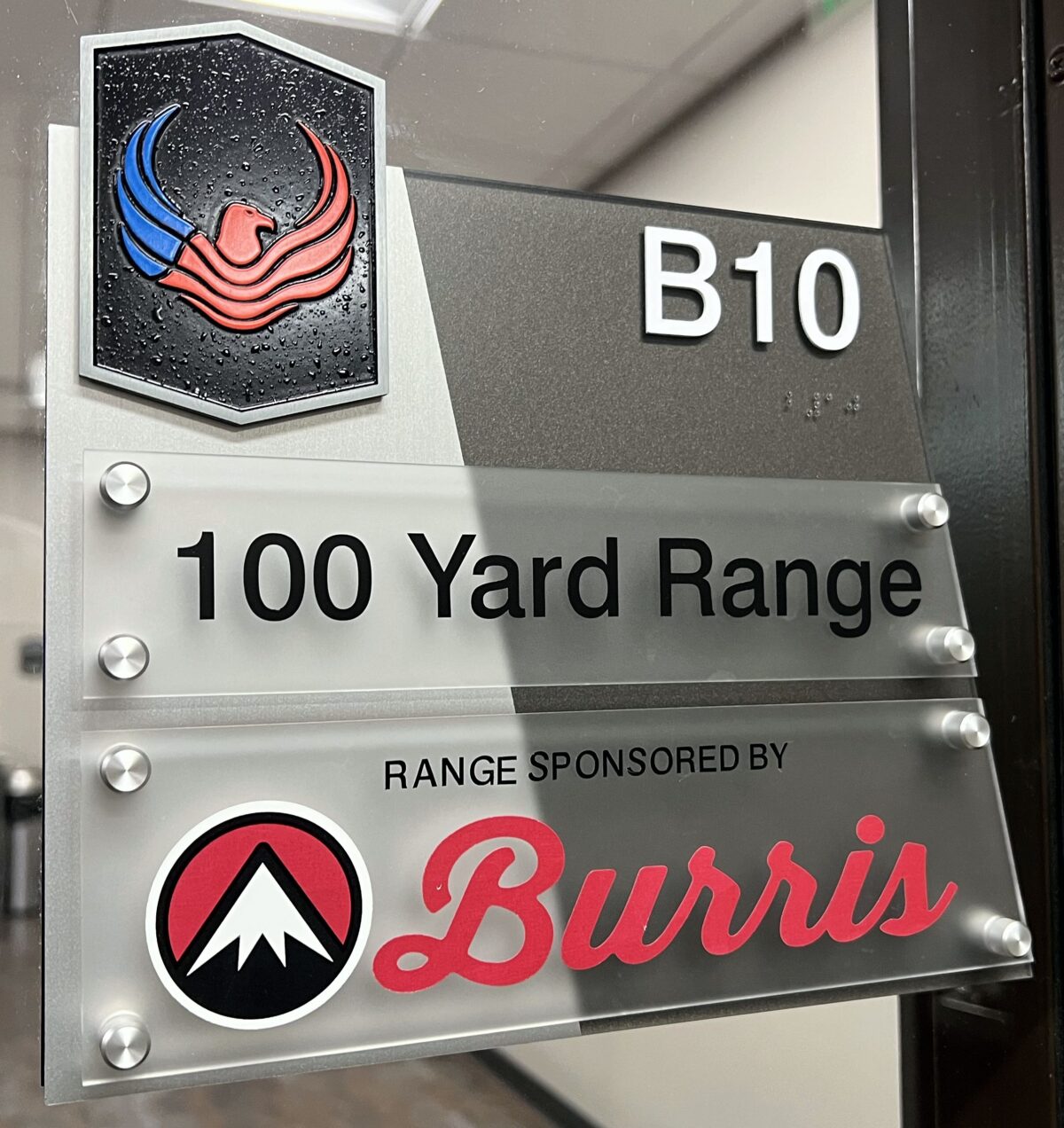 A sign with a black and red phoenix logo at the top, labeled "B10". Below it reads "100 Yard Range". The bottom part of the sign states "Range Sponsored by Burris" with the Burris company logo.