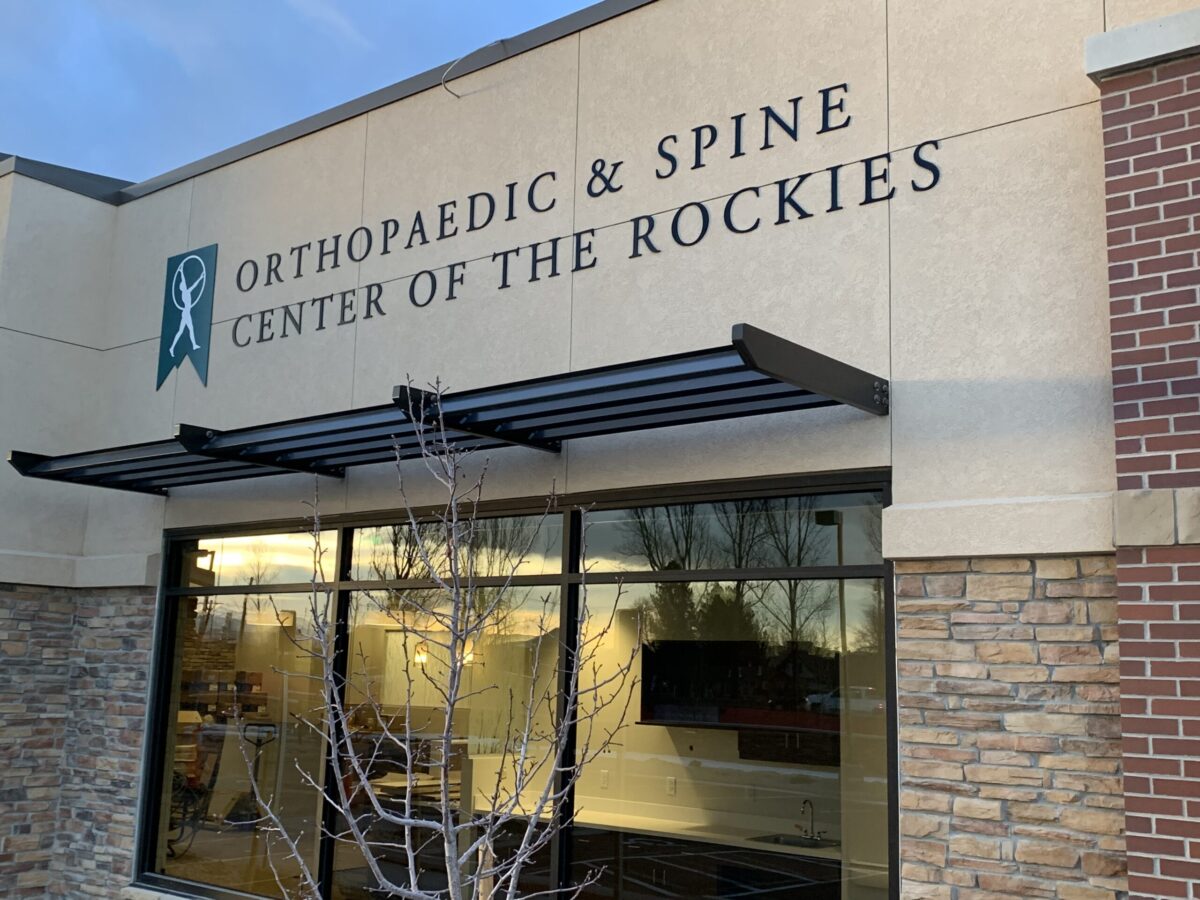 The image shows the exterior of the Orthopaedic & Spine Center of the Rockies building. The signage above the entrance features the center's name in large, dark letters. The building exterior includes stonework, large windows, and a small awning.