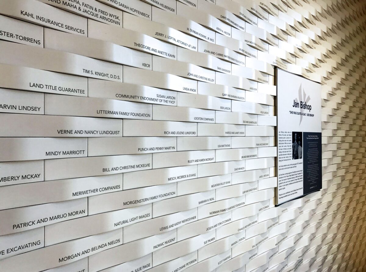 A wall is covered with rows of rectangular white plaques, each bearing different names and messages. To the right, a larger black and white informational plaque is visible. The pattern of the wall creates a textured, wave-like visual effect.