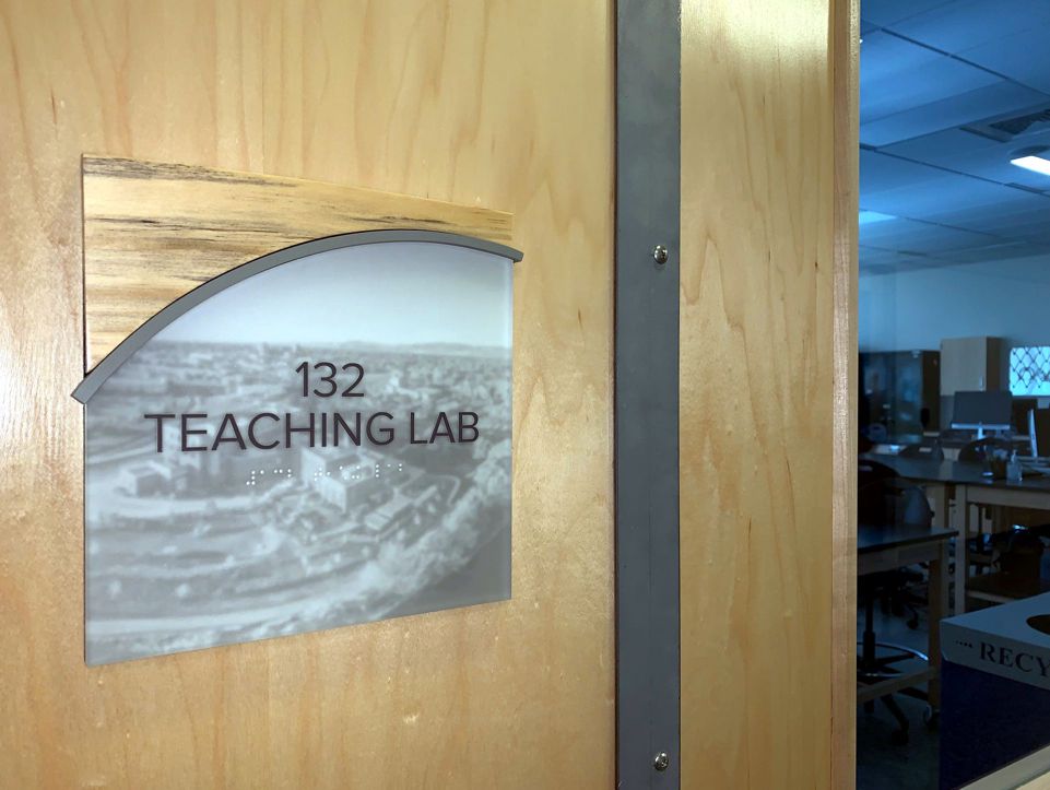 A wooden door with a sign that reads "132 Teaching Lab" in black letters over a black and white photo.