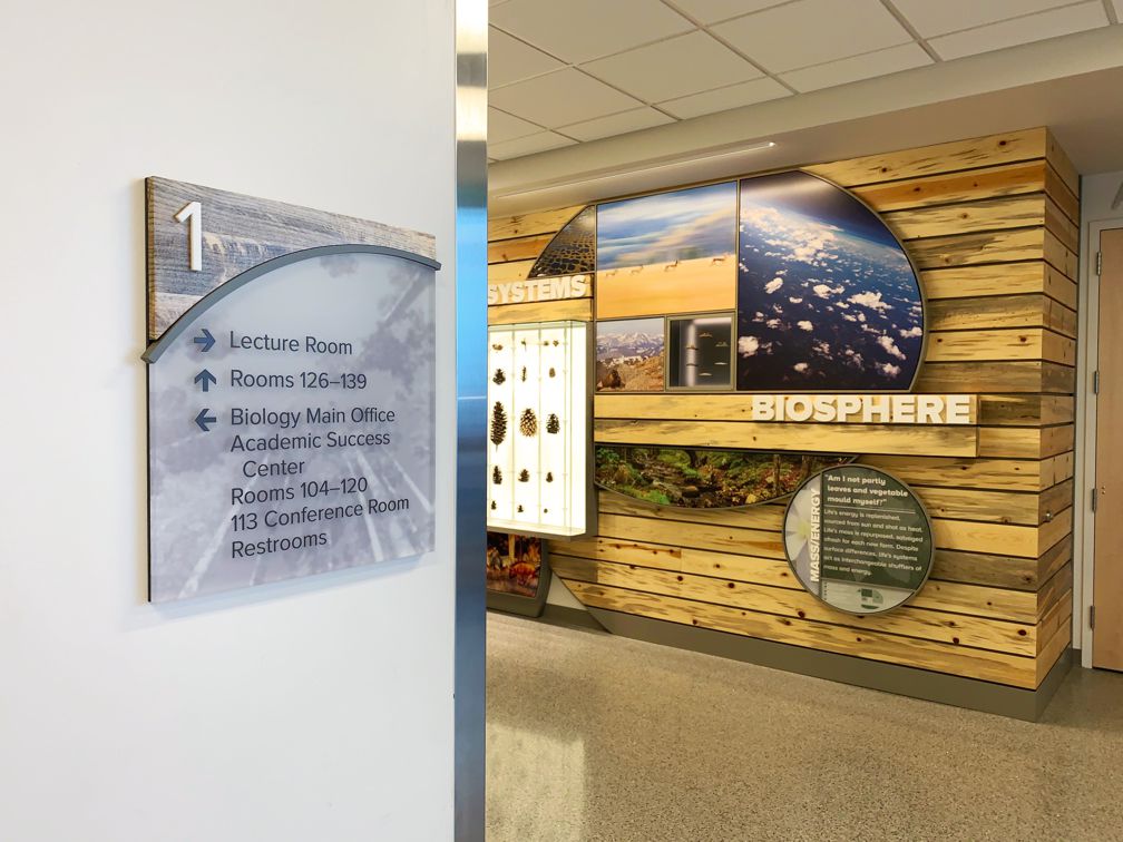 A wall sign on the first floor directs to a lecture room, rooms 126-139, the Biology Main Office, Academic Success Center, rooms 104-120, and a conference room (113). The sign uses a mix of blue and black text with arrows beside each listing.