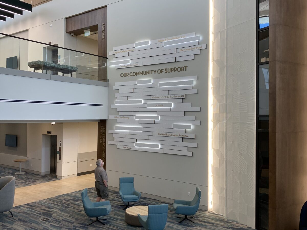A modern indoor space featuring a high ceiling and contemporary decor. On one wall, there is a large, artistic installation with the text "Our Community of Support." Several chairs and a round table are arranged on the floor.