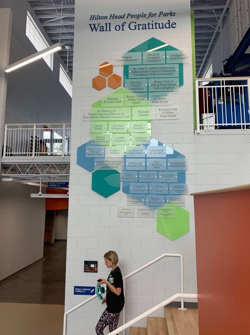 A woman walks up the stairs inside a modern building, passing by a "Wall of Gratitude" display on a large white wall. The display features colorful hexagon shapes containing names and text, signifying recognition and appreciation.