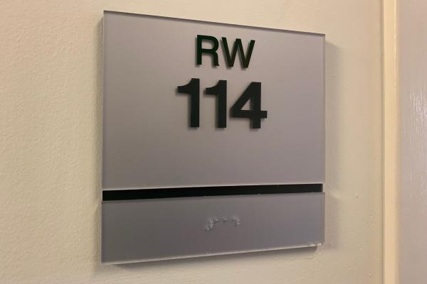 A sleek, grey wall sign with bold green lettering reading 'RW 114', featuring Braille underneath.