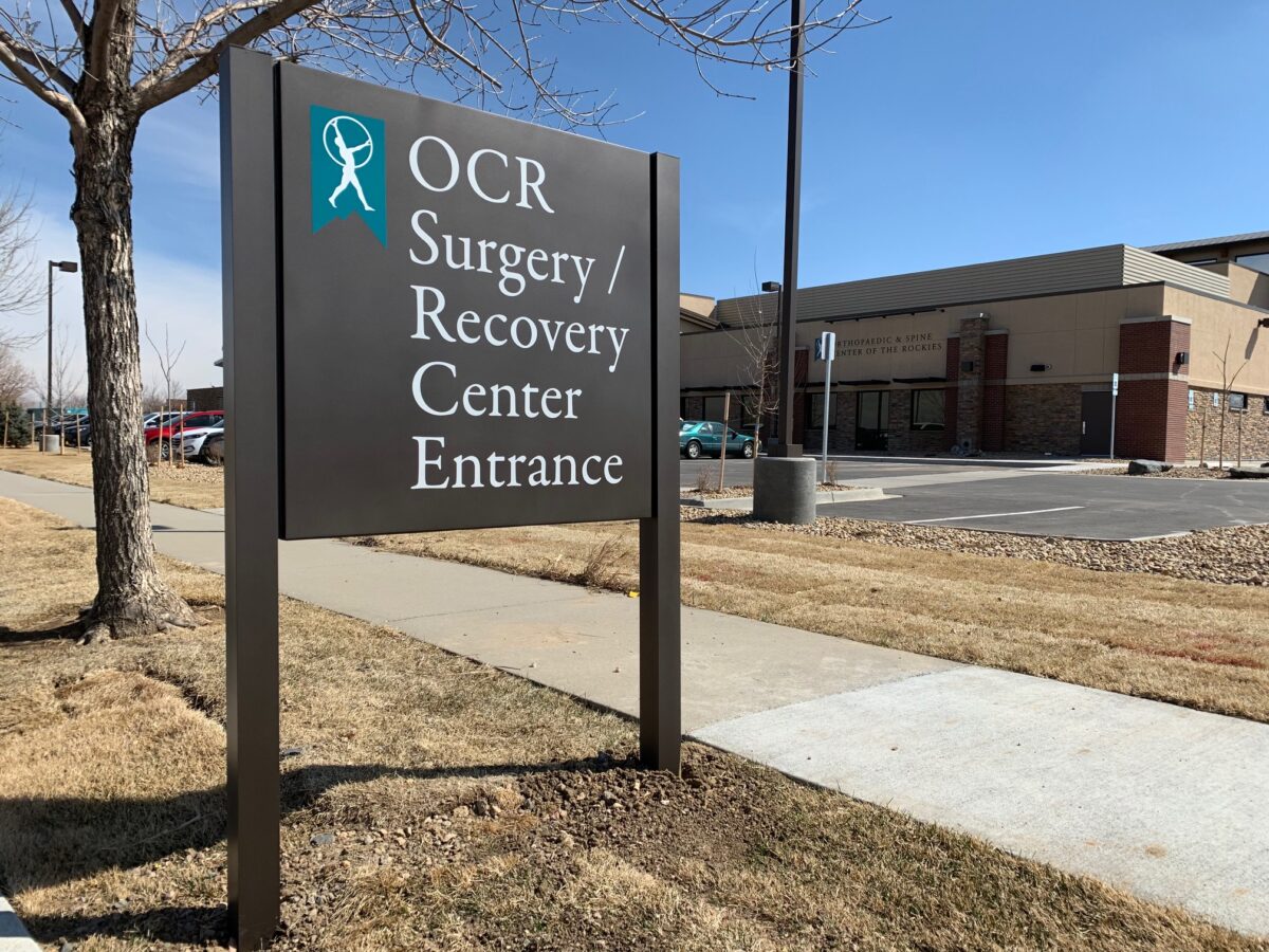 Sign near a sidewalk reads "OCR Surgery / Recovery Center Entrance" with a teal and white logo above the text. A building and parking lot are visible in the background. It is a sunny day with a clear sky.