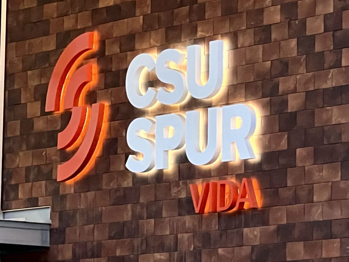 The image shows a wall sign with the text "CSU SPUR VIDA" illuminated. The letters "CSU" and "SPUR" are in white, while the word "VIDA" is in red. The wall is made of brown rectangular tiles, and the letters are outlined with a glowing effect.