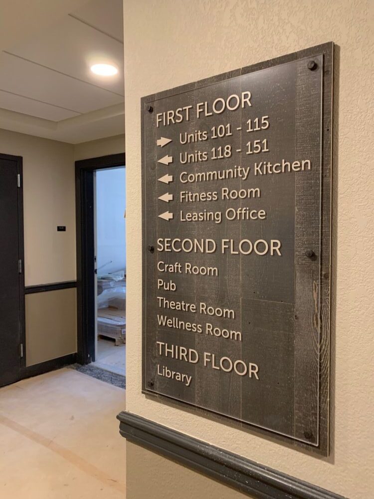 A directional sign mounted on a wall inside a building lists the locations of various rooms and facilities. A hallway and door are visible to the left.