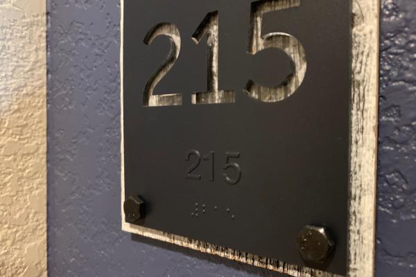 This image displays a room number sign, "215," which is crafted with raised silver numerals on a distressed black background, held in place by industrial-style rivets.