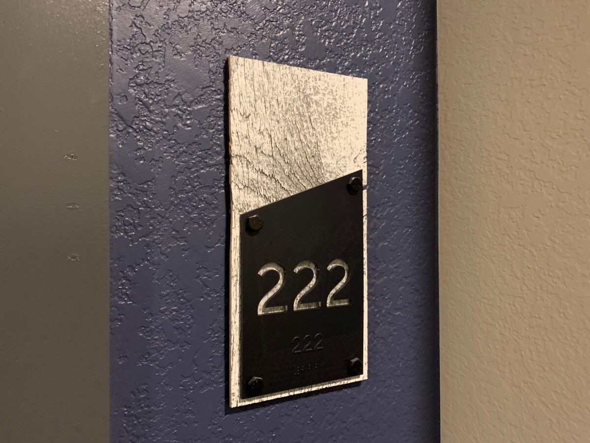 A room number plaque mounted on a textured wall. The plaque has a metallic background with a black section displaying the number "222" in raised text. The wall behind the plaque is painted dark blue.