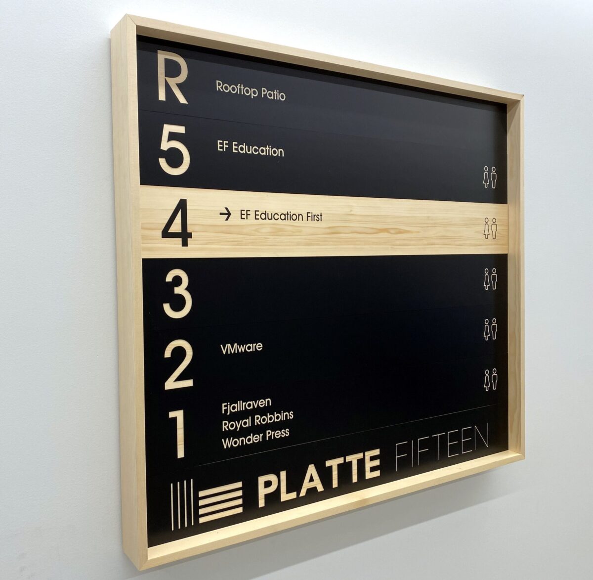 A rectangular directory sign with a wooden frame is mounted on a wall. It lists floors and corresponding occupants: 5 - Rooftop Patio, EF Education; 4 - EF Education First; 3 - VMware; 2 - Fjallraven, Royal Robbins, Wonder Press. The bottom reads "PLATTE FIFTEEN.