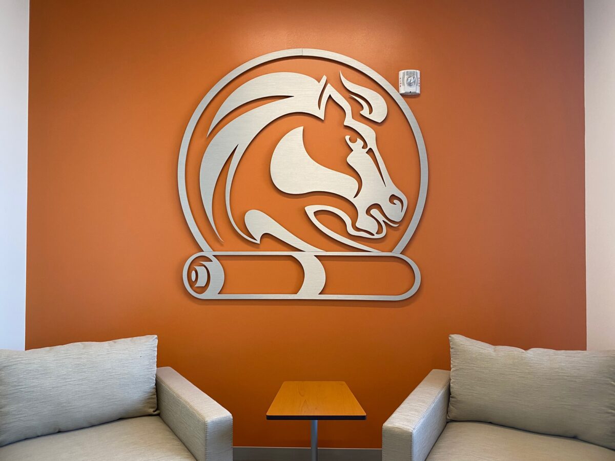A wall adorned with an orange matte finish displays a stylized horse logo above a scroll. The logo is prominently centered. In front of the wall, two cushioned chairs in a light fabric face each other with a small wooden table placed between them.