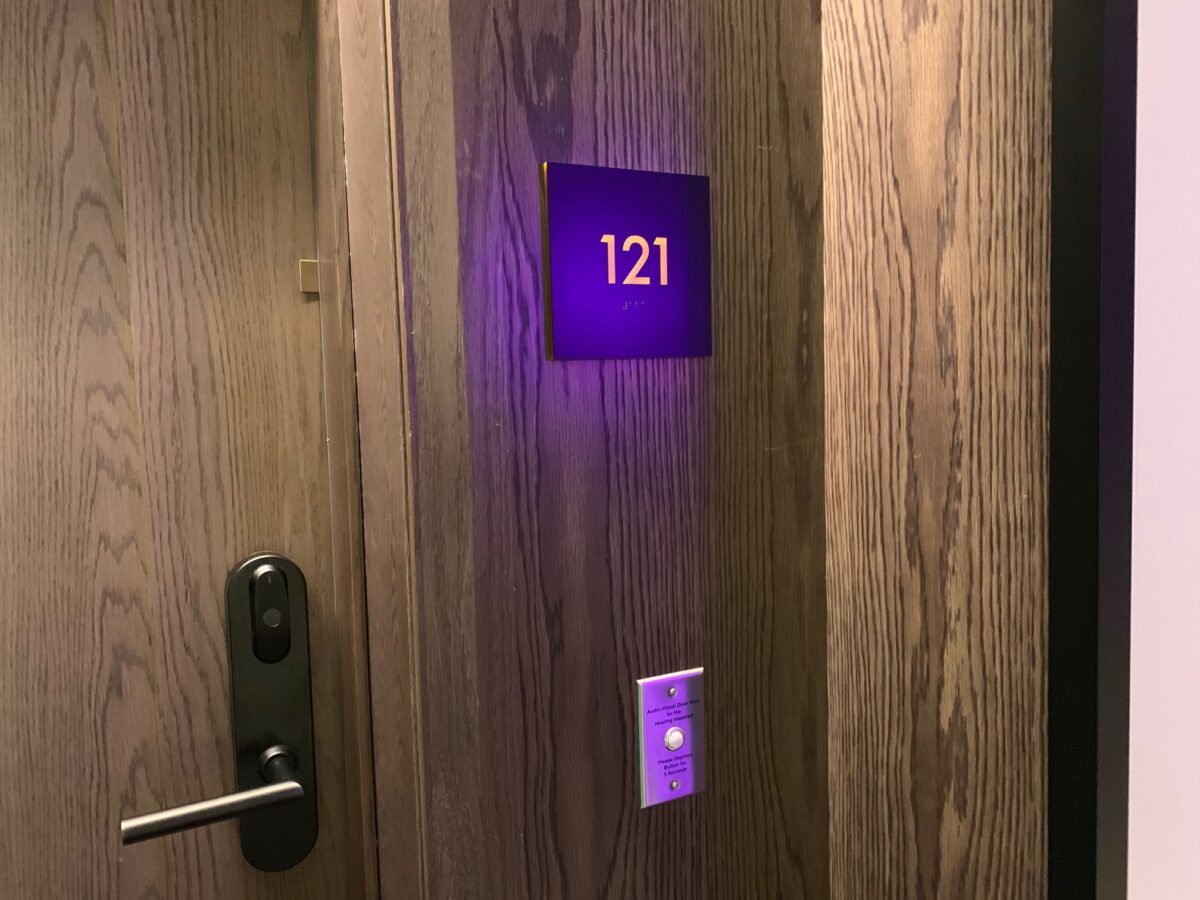 A hotel room door with the number 121 illuminated in purple on a sign above the door handle. The door is made of wood, with a sleek black electronic lock and key card reader below the door number.
