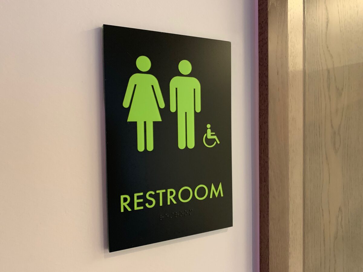 A restroom sign showing green, simplified icons for male, female, and accessible restrooms on a black background. The sign is mounted on a wall next to a wooden door. The word "RESTROOM" is written in green letters beneath the icons.