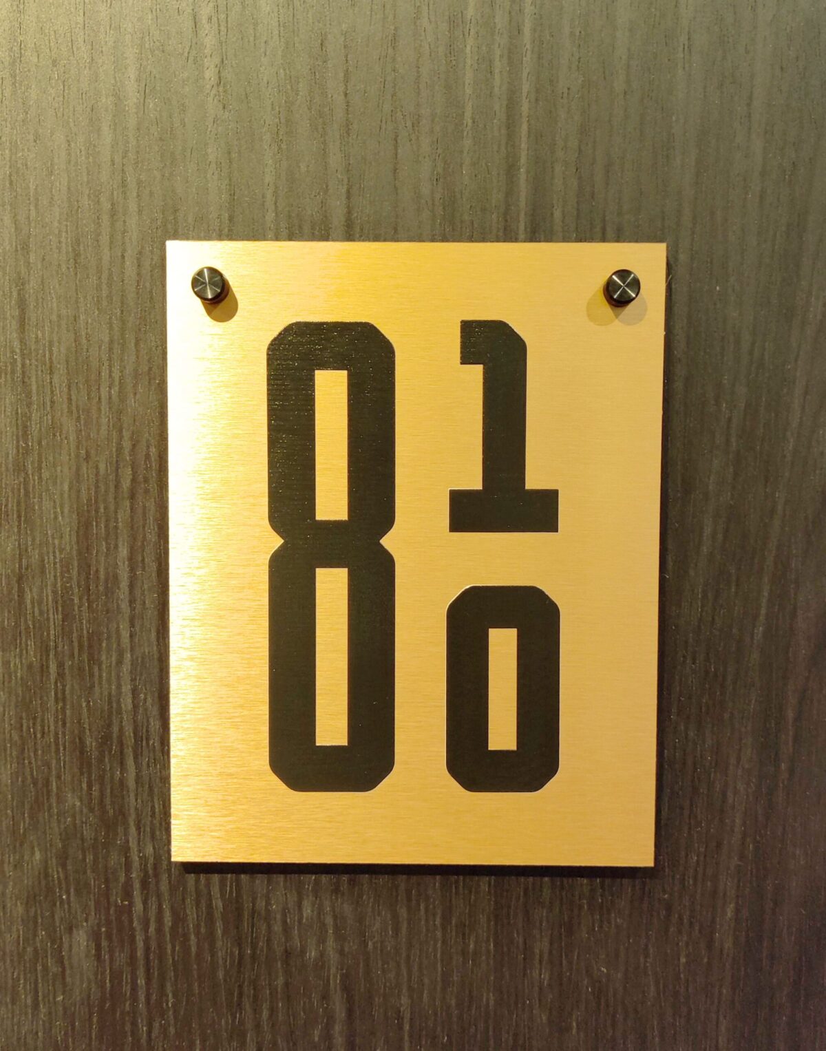 A gold rectangular plaque mounted on a dark wooden surface with the numbers "810" in large black font. The plaque is held in place by two screws, one at each upper corner.