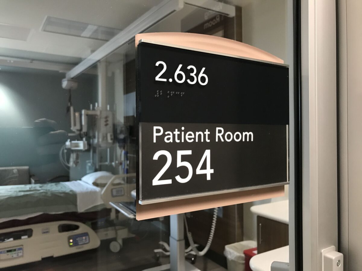 A hospital room sign indicates "2.636" and "Patient Room 254" in white text on a black background. The room, visible through a glass window, contains medical equipment and a bed with white sheets.