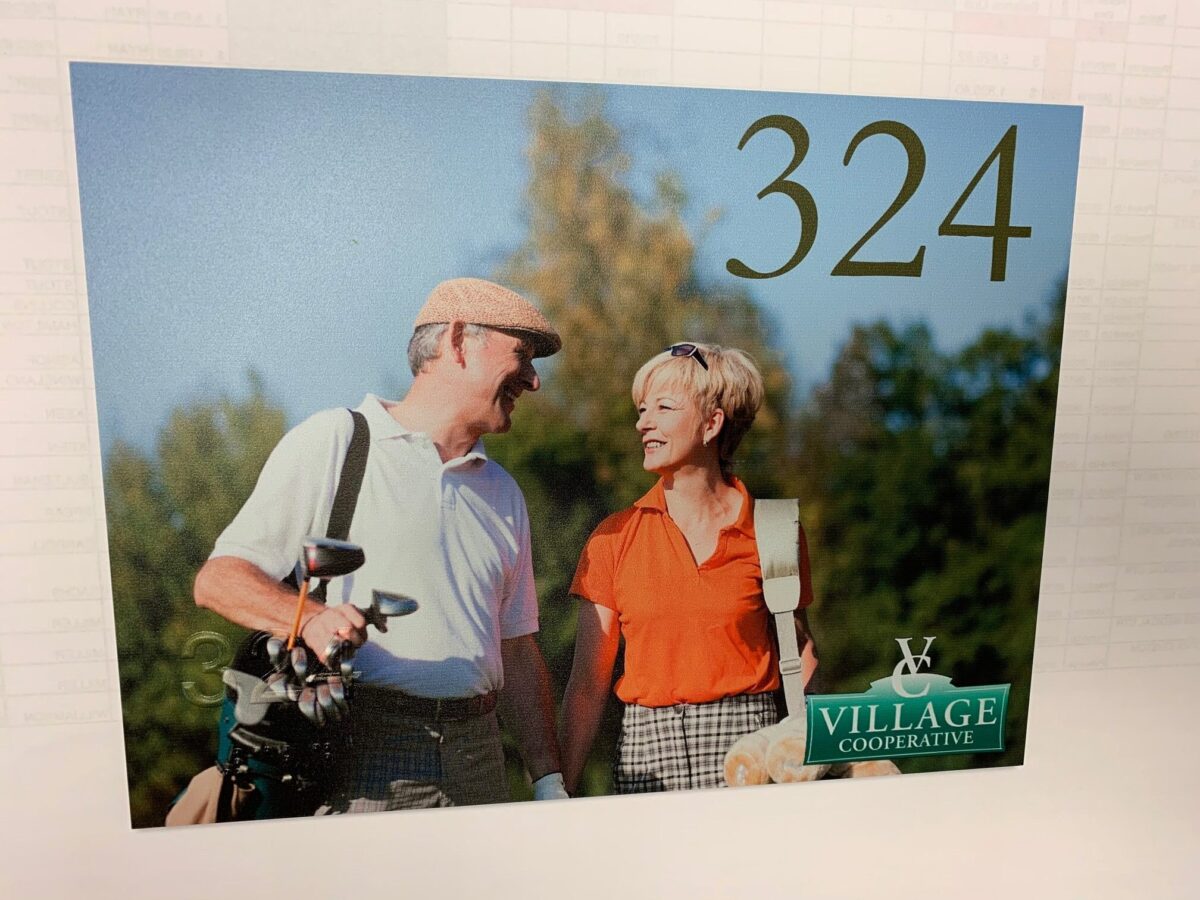 A man and a woman, both smiling, stand outdoors under a clear blue sky. The man wears a cap, holds a golf bag, and the woman is in an orange shirt. They appear to be on a golf course. The number "324" is in the top right corner, with a "Village Cooperative" logo in the bottom right, and braille writing in the bottom left.