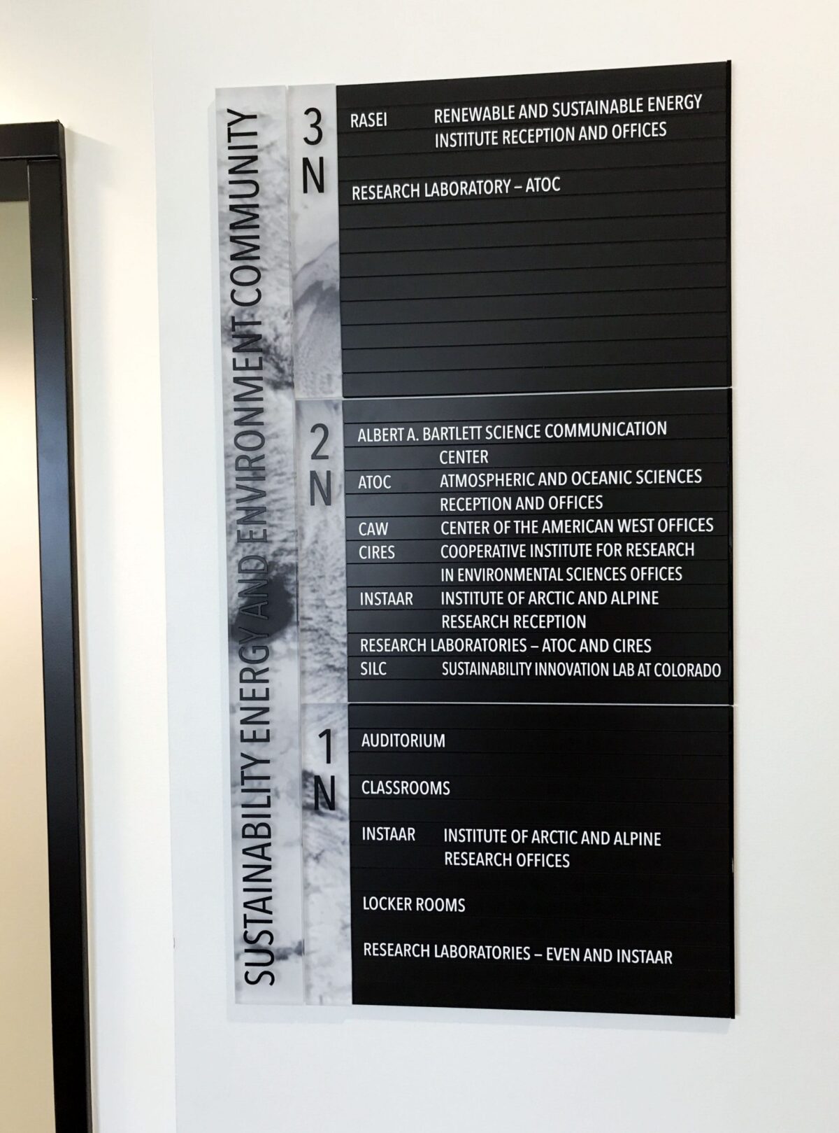 A wall-mounted black and white office directory sign labeled "Sustainability Energy and Environment Community." The sign shows room numbers and corresponding offices or departments. The details of the text are mostly blurry and not fully readable.